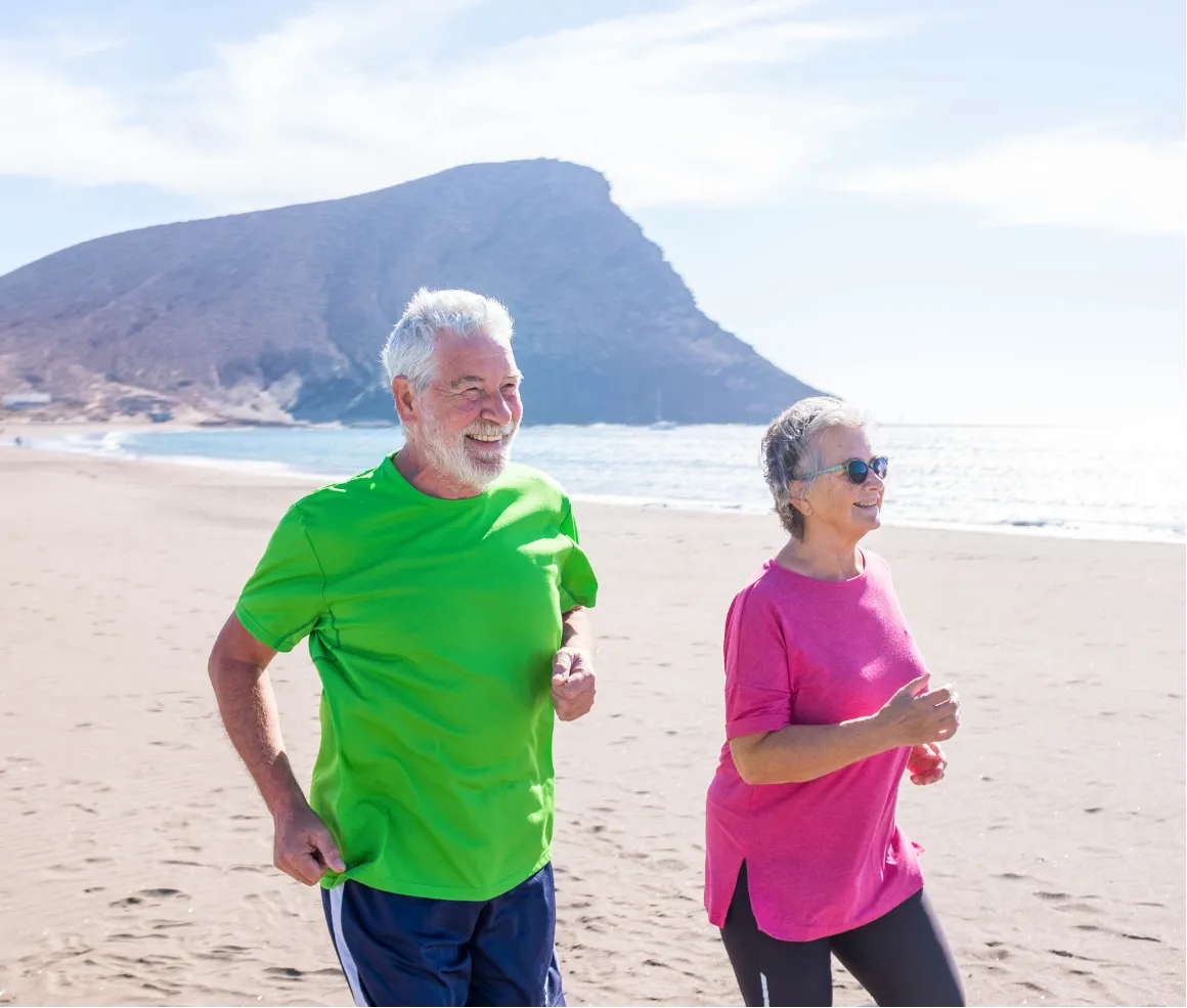 Two older adults jogging on a sandy beach with a mountain and ocean in the background on a sunny day.