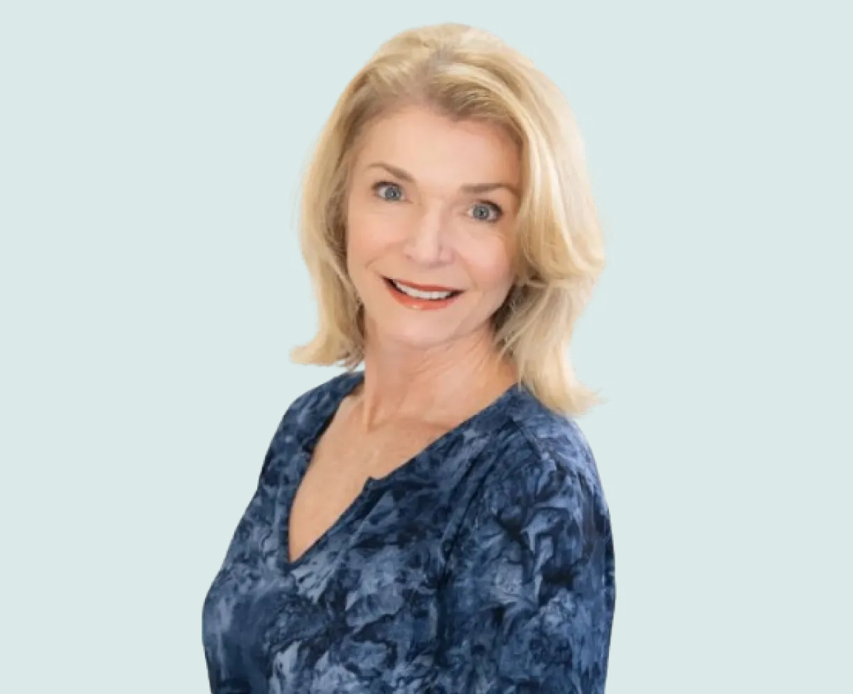 Smiling older woman with blonde hair wearing a blue patterned top on a teal background.