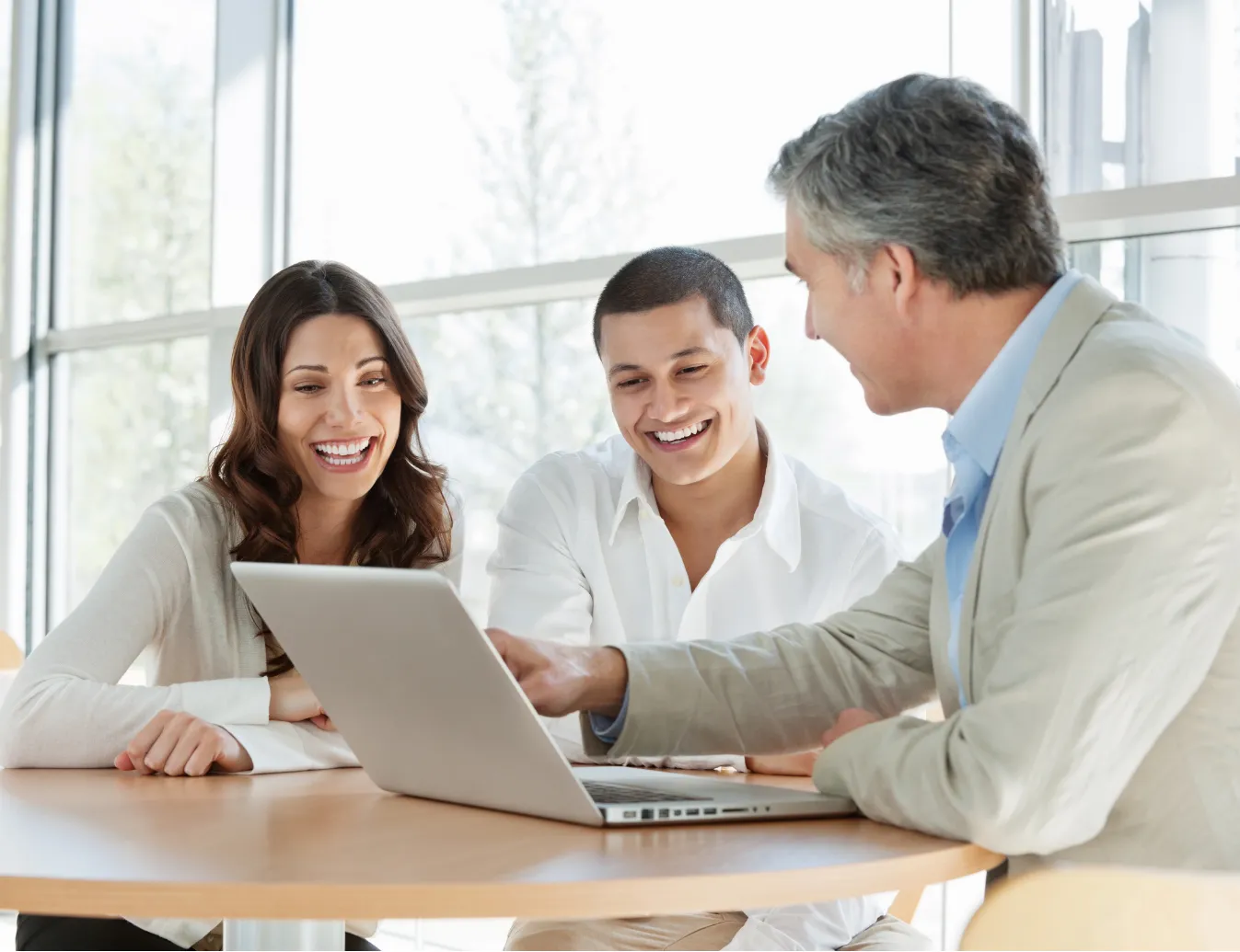Three people sit at a table, smiling and looking at a laptop in a bright office setting.
