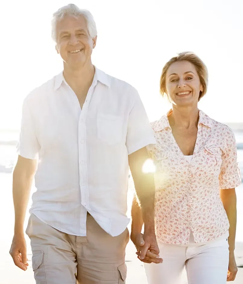 Smiling older couple holding hands and walking on a sunny beach, dressed in light, casual clothing.