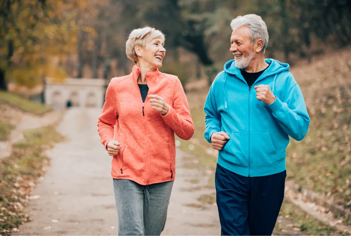 Older couple jogging together outside on a park path, smiling and wearing sporty jackets.