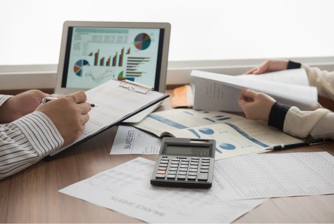 Two people review financial documents at a desk with a laptop showing charts and a calculator nearby.