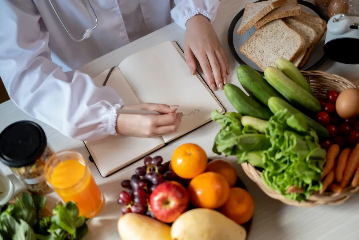 A person writes in a notebook near fresh fruits, vegetables, juice, and bread on a table.