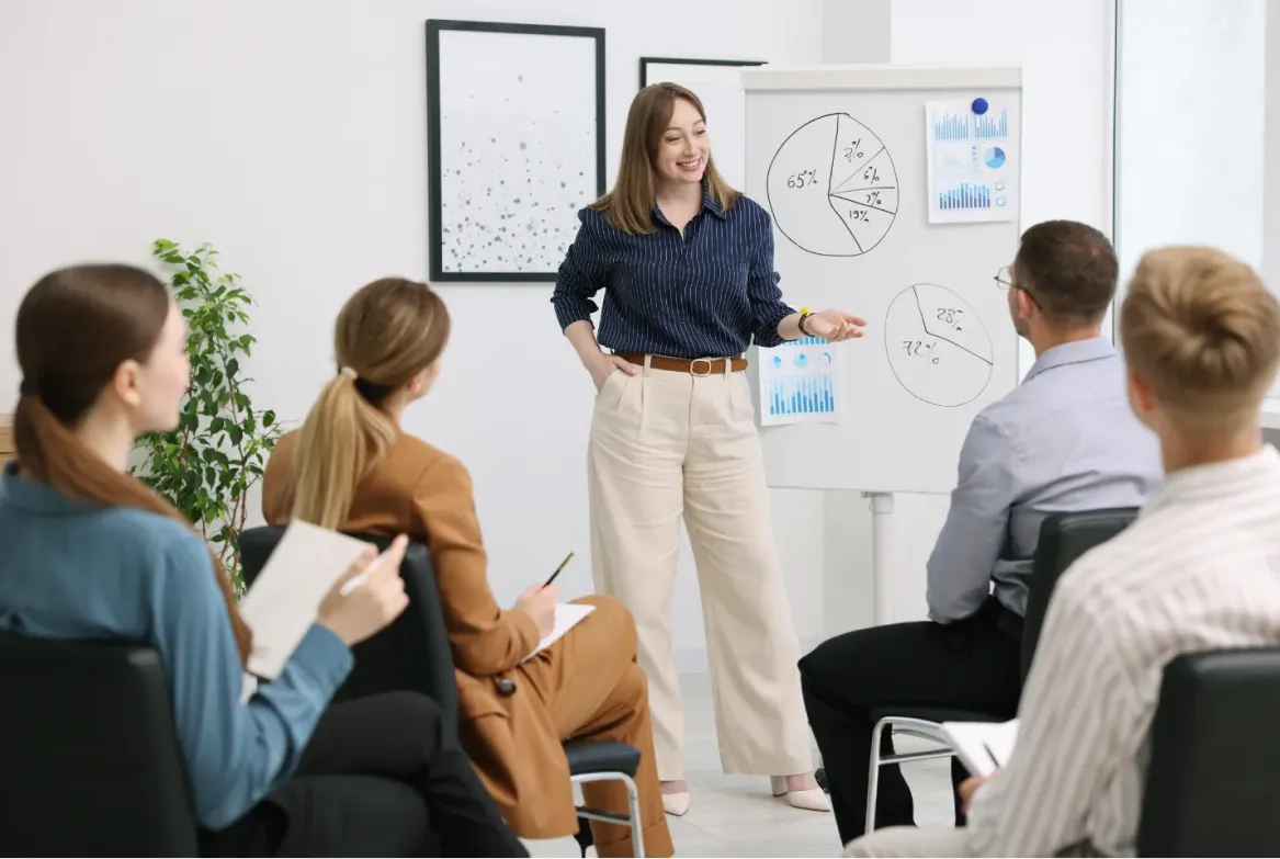 Woman presents charts to seated group in office setting; attendees listen and take notes.