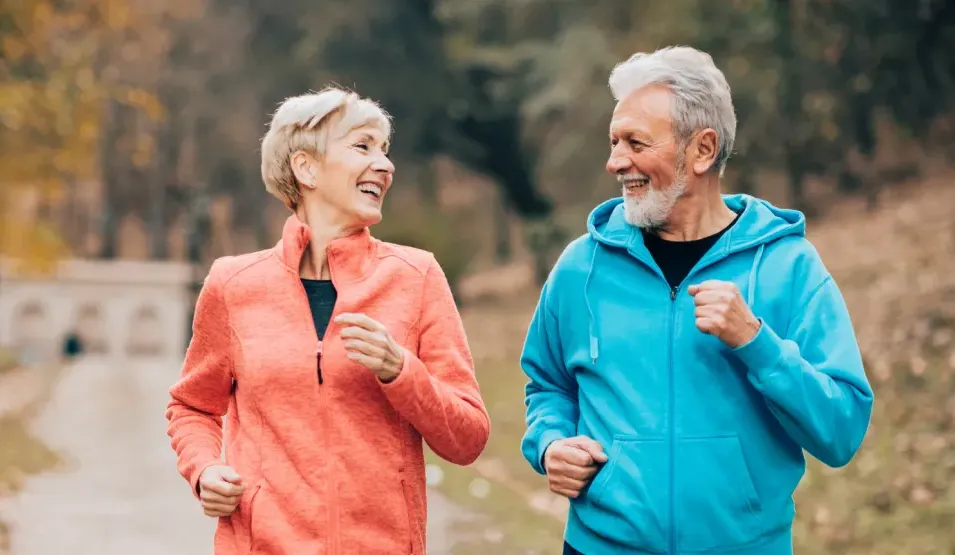 Two smiling older adults in athletic wear jogging together outdoors on a tree-lined path.