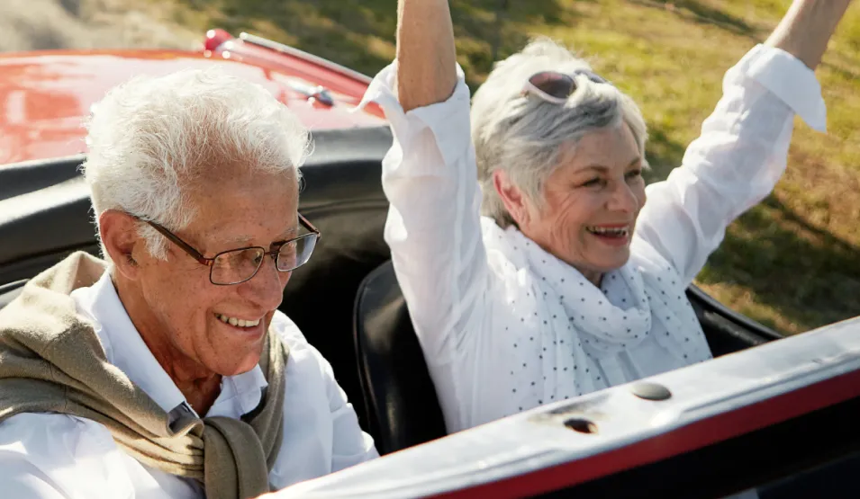 Smiling older couple enjoys a convertible car ride; woman raises her arms joyfully, both wear glasses and light clothes.
