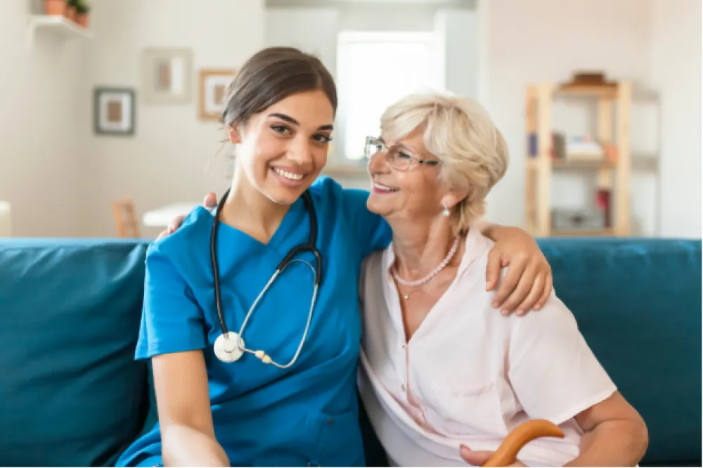 Young nurse in blue scrubs sitting on a couch, smiling and hugging an elderly woman with glasses and white hair.