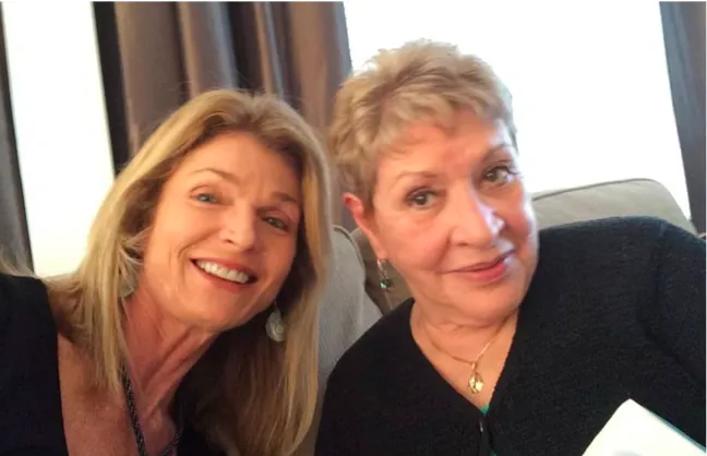 Two women smiling and sitting together indoors, one taking a selfie while the other looks at the camera.