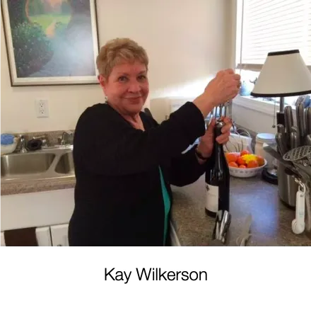 Woman in a kitchen smiling, opening a wine bottle with a corkscrew, fruit on the counter nearby.